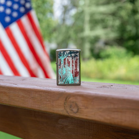 Lifestyle image of Statue Of Liberty Design Black Ice® Windproof Lighter standing on a railing with an American Flag in the background.