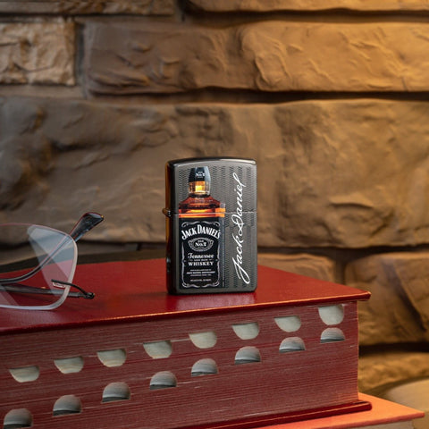 Lifestyle image of Jack Daniel's® Logo and Bottle Gray Windproof Lighter standing on a red book with glasses next to it